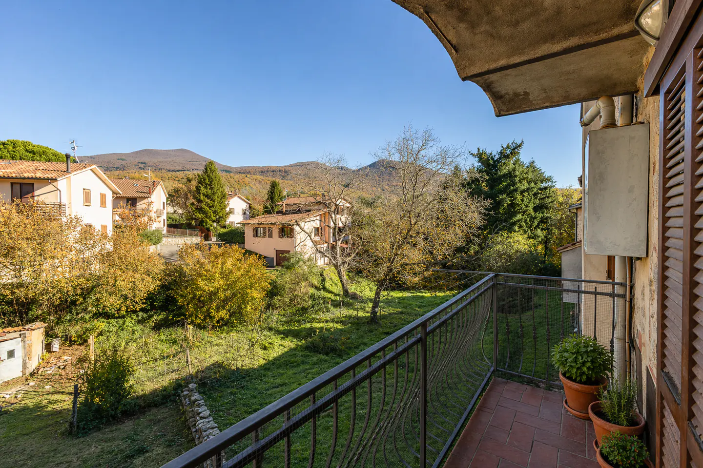 View from a balcony with a black metal railing, overlooking a green lawn, houses, trees, and mountains under a blue sky. Potted plants sit on the red-tiled balcony.