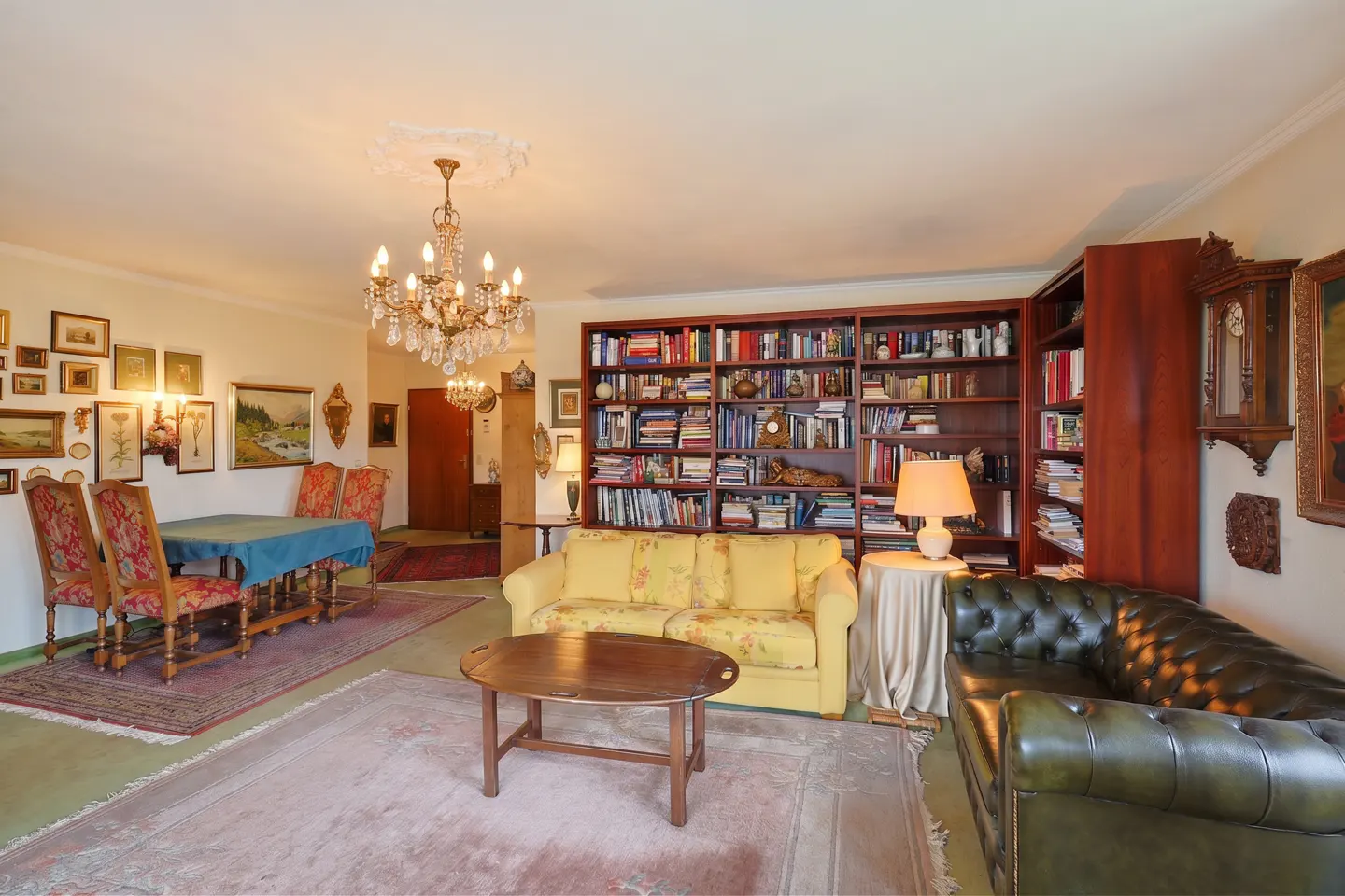 Living room with a yellow sofa, green leather couch, wooden coffee table, dining area, and large floor-to-ceiling bookshelf.