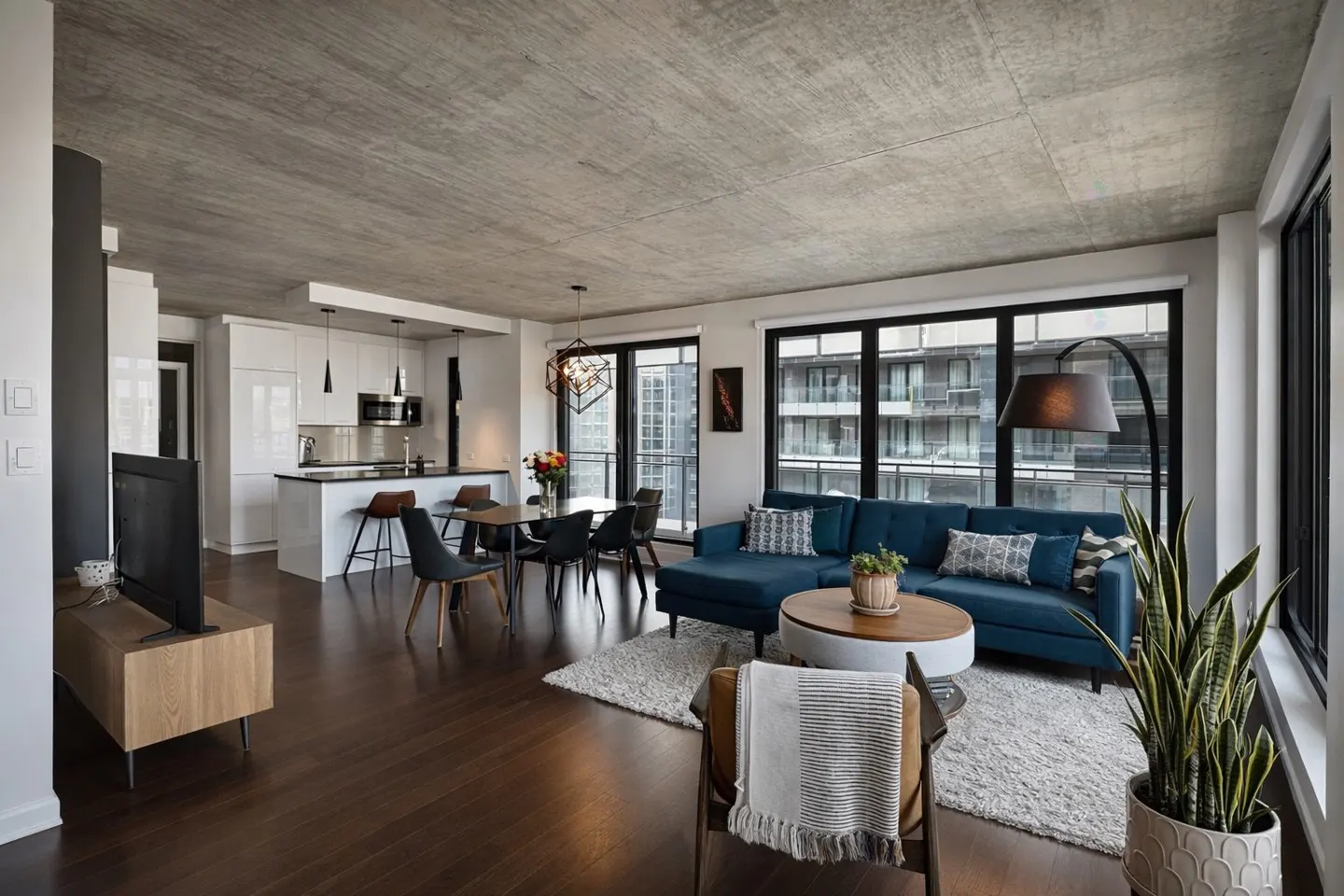 Open-concept apartment with a blue sofa, dining table, and white kitchen. Large windows offer city views.