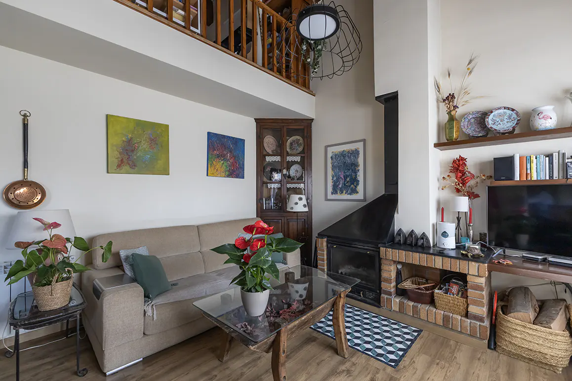 A living room with a beige sofa, glass coffee table, fireplace, and a loft above. Artwork adorns the walls, and plants add a touch of nature.