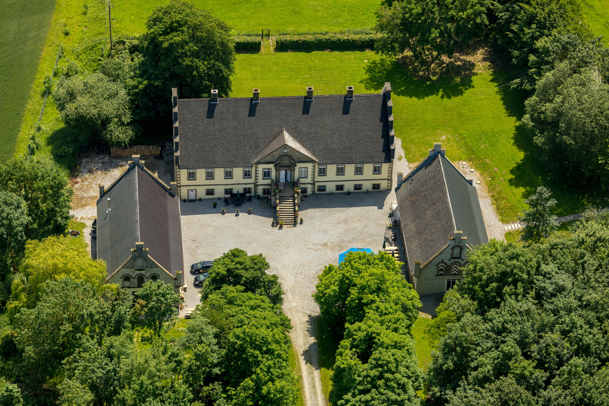 Aerial view of a large, light yellow manor house with a dark roof, surrounded by green trees and lawns.