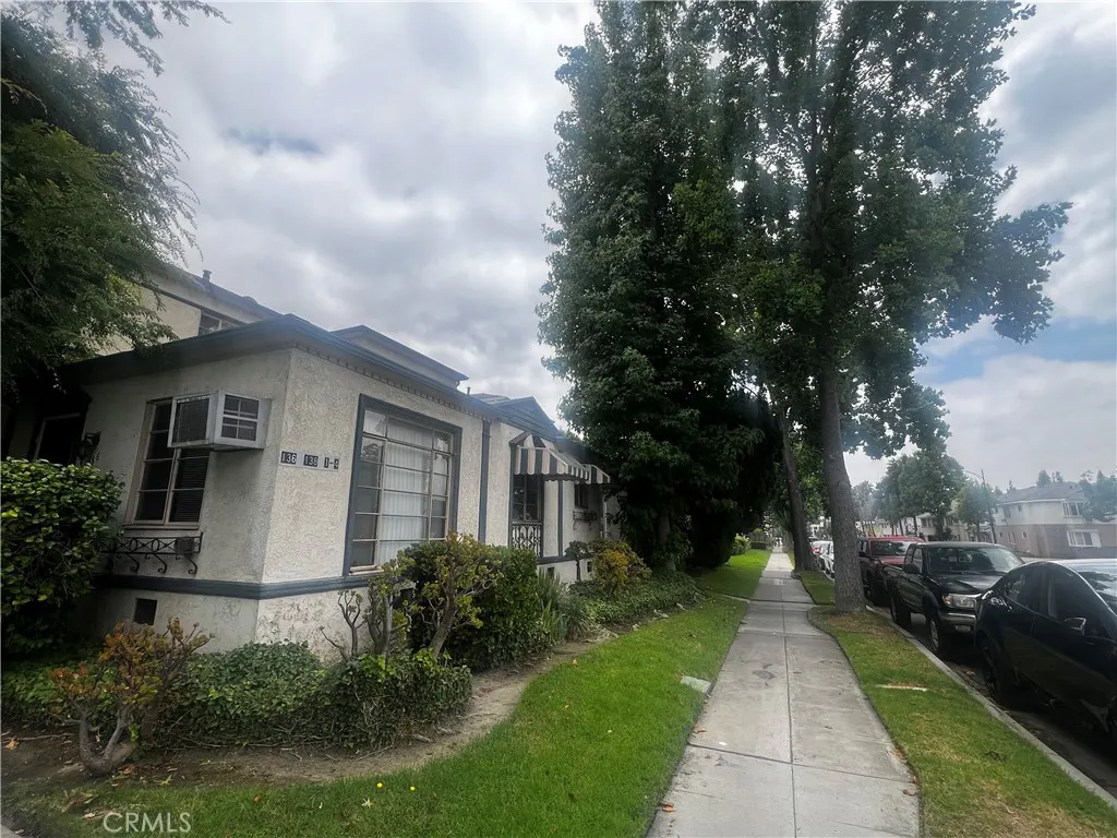 Exterior view of a light beige stucco apartment building with dark trim, green bushes, and a sidewalk. Cars are parked along the street.