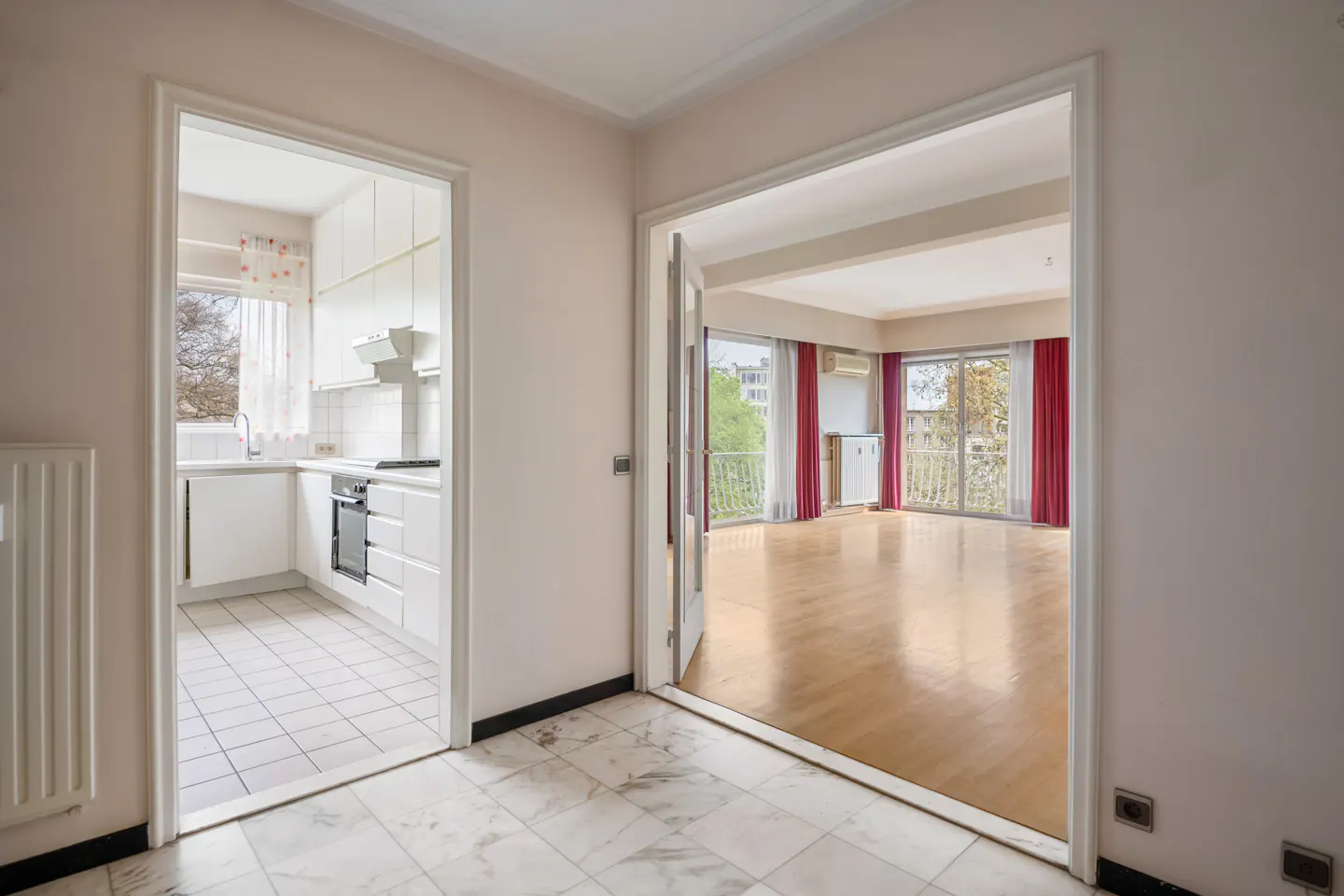 A view of a kitchen and living room through doorways. The kitchen has white cabinets and appliances. The living room has wood floors and red curtains.