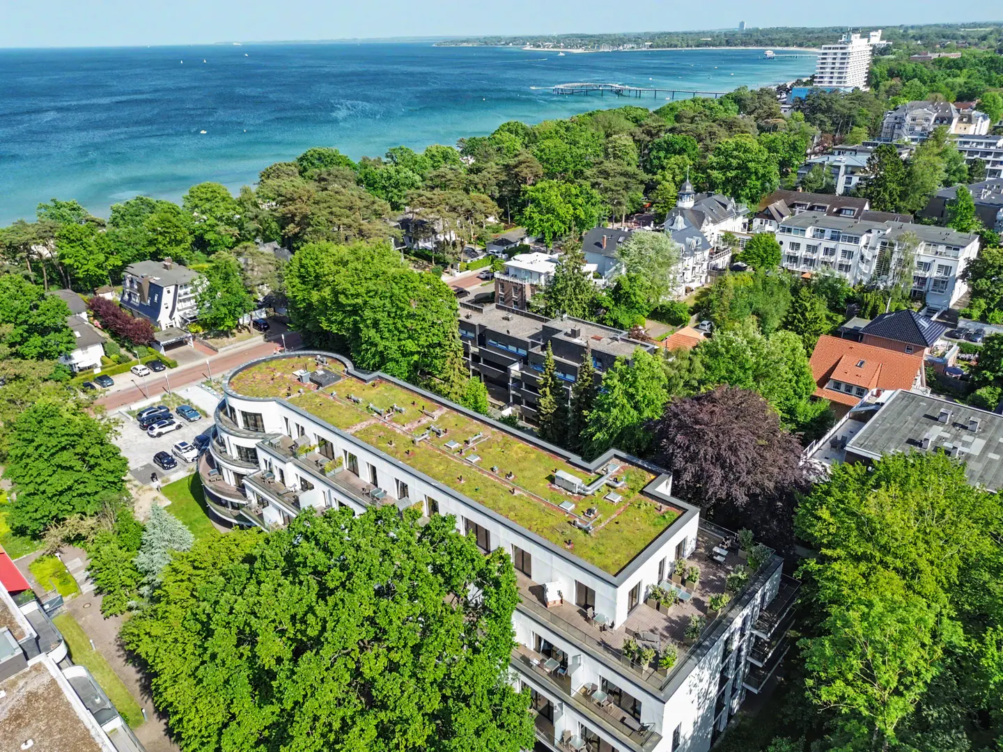 Aerial view of a modern white apartment building with a green roof, surrounded by trees near a blue coastal bay.