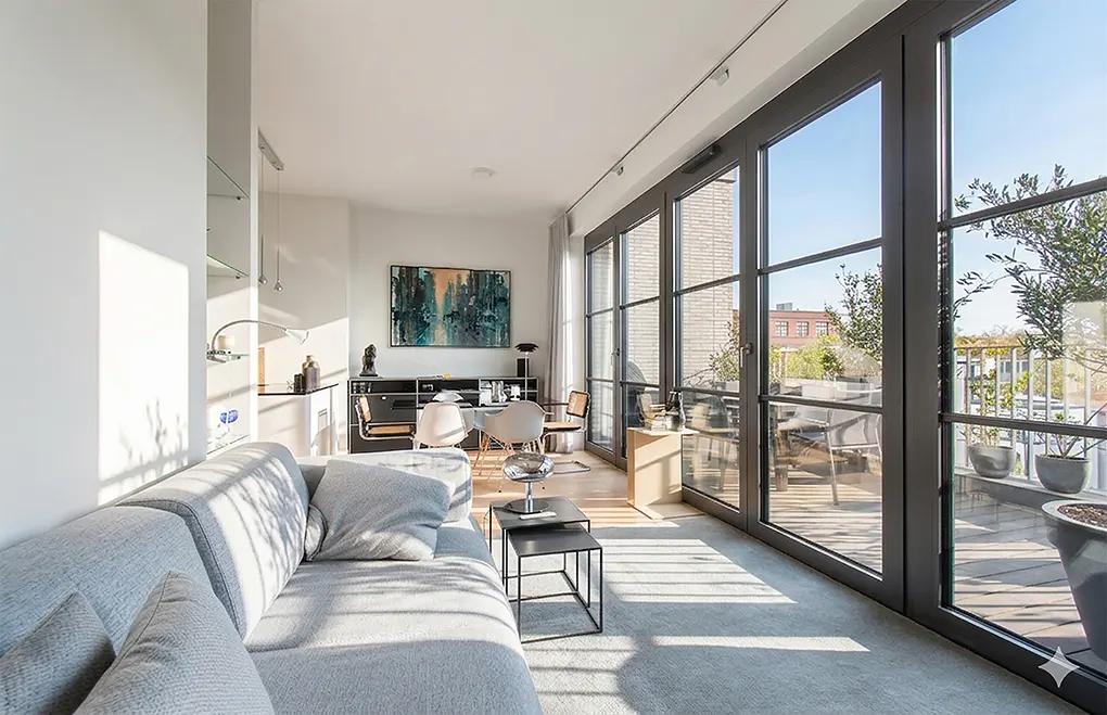 Bright, modern living room with a gray sofa, black tables, and a dining area. Large windows open to a balcony with plants.