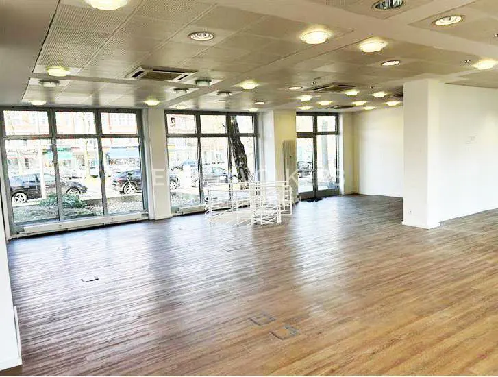 Bright, empty commercial space with wood floors, white walls, and large windows. Cars are visible outside.