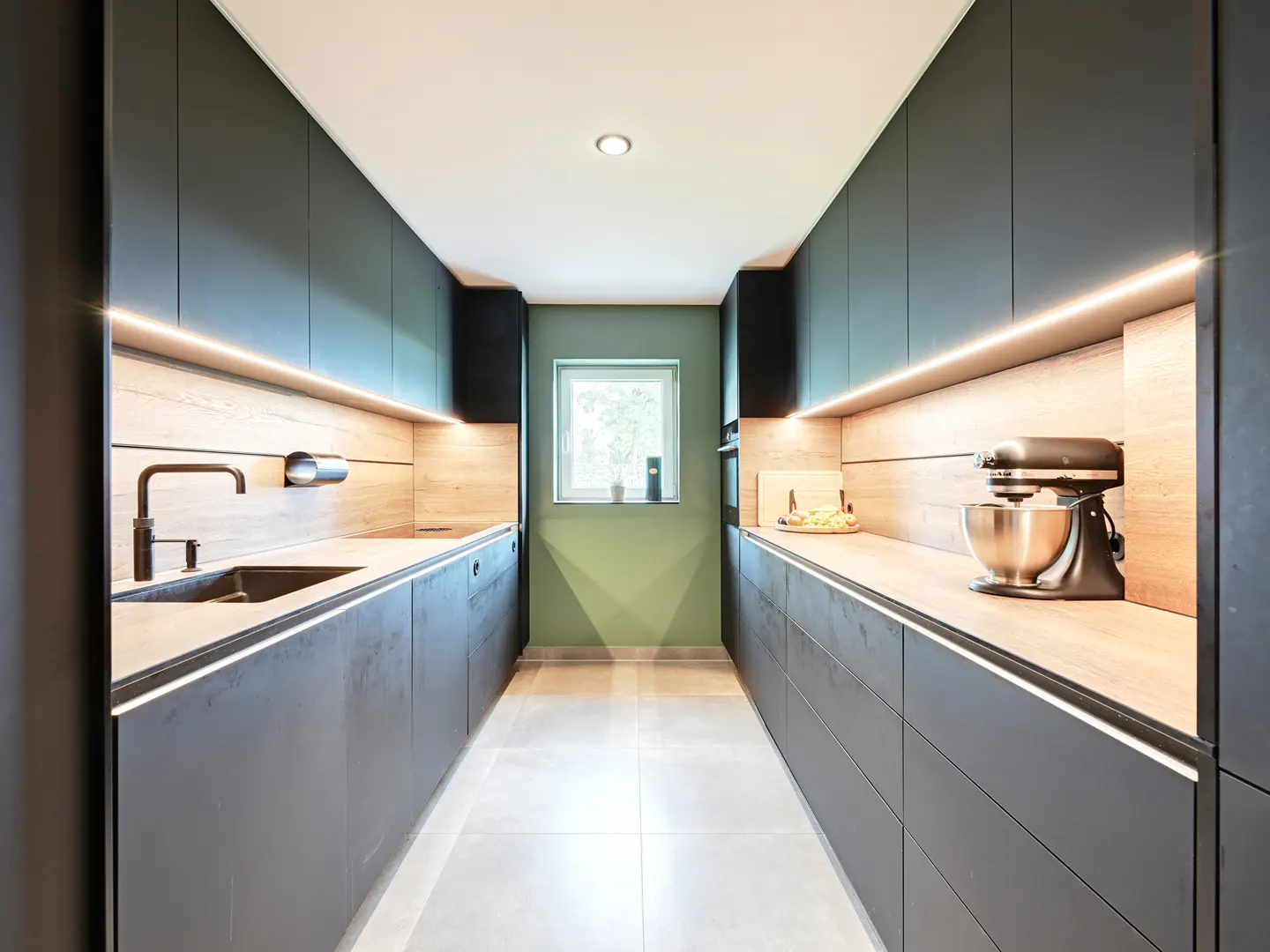 A modern kitchen with gray cabinets, wood countertops, and LED strip lighting. A window is centered at the end of the galley kitchen.