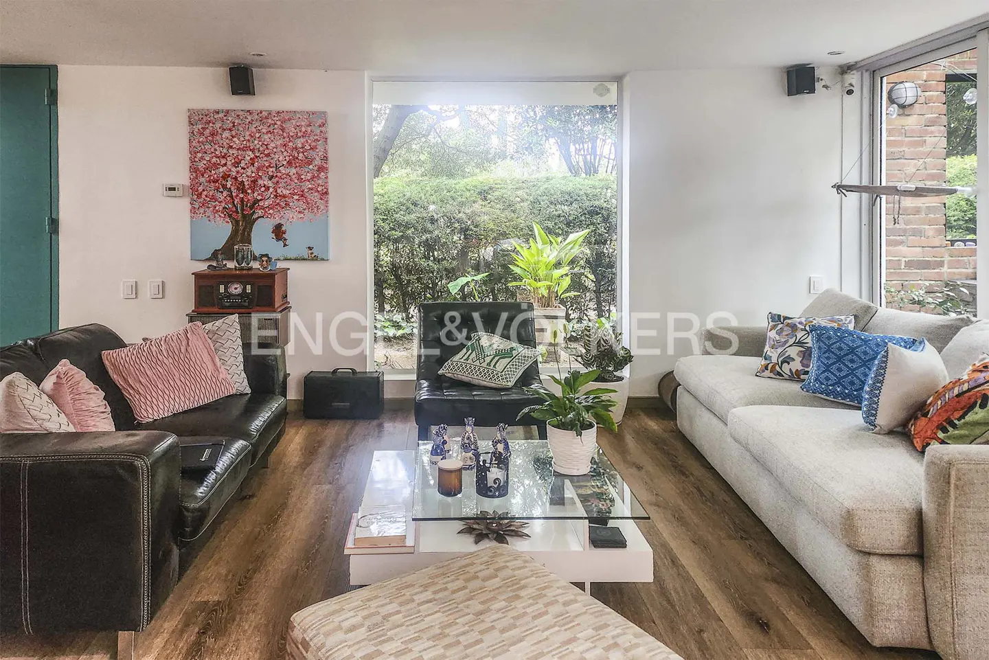 Living room with a black leather sofa, a light gray sofa, a glass coffee table, and a large window overlooking a garden. A painting of a pink tree hangs on the wall.