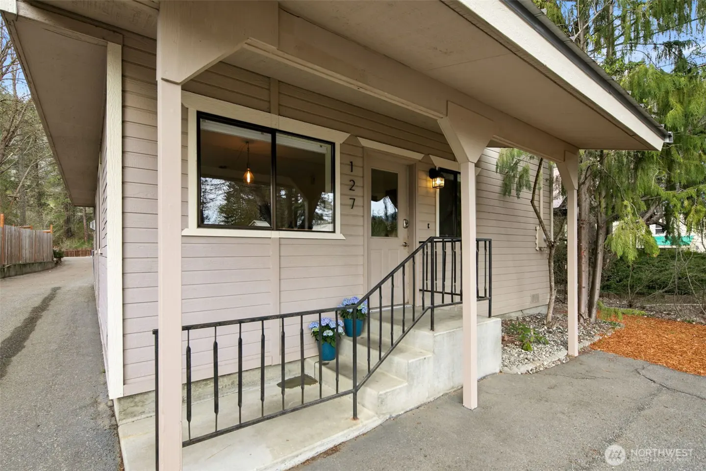 Exterior view of a beige house with a covered porch, black railing, and concrete steps. Blue flowers in pots adorn the steps. The house number "127" is visible above the door.
