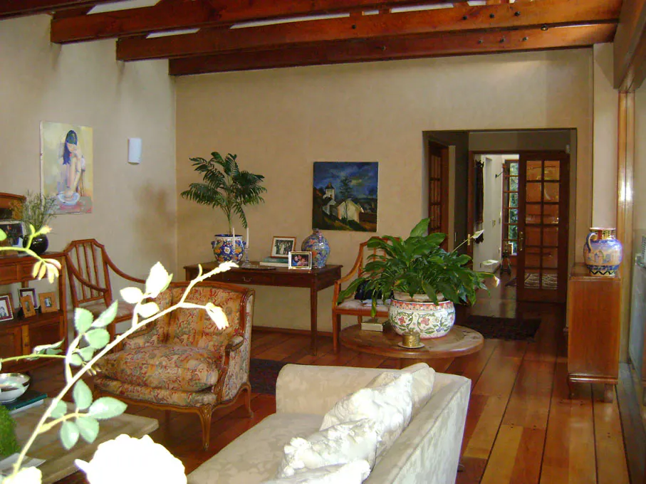 Living room with wood floors, exposed beams, and tan walls. Furniture includes a cream sofa, patterned chairs, and wooden tables. Paintings and plants add color.