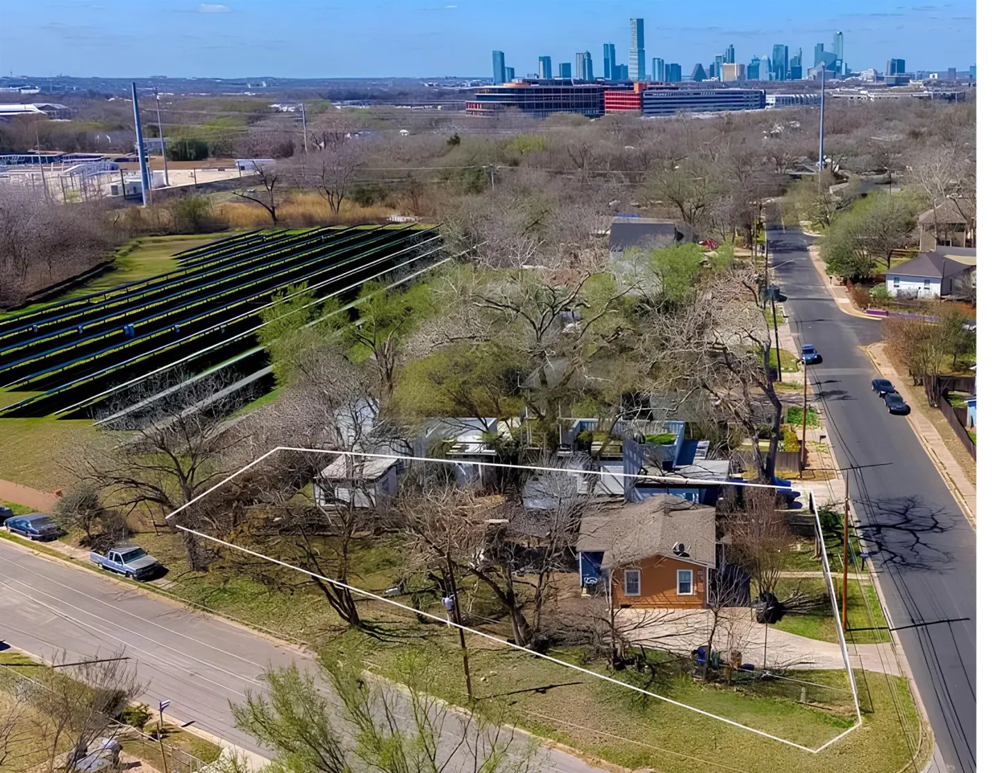 Aerial view of a property outlined in white, featuring an orange house, trees, and a street with cars. A solar panel field and city skyline are in the background.