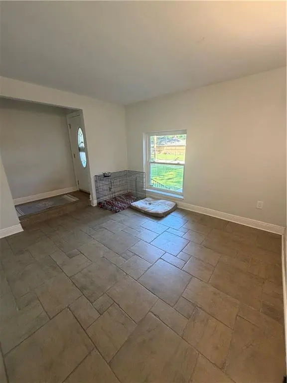 A bright, empty living room with beige tile flooring, white walls, a window, and a dog crate and bed.