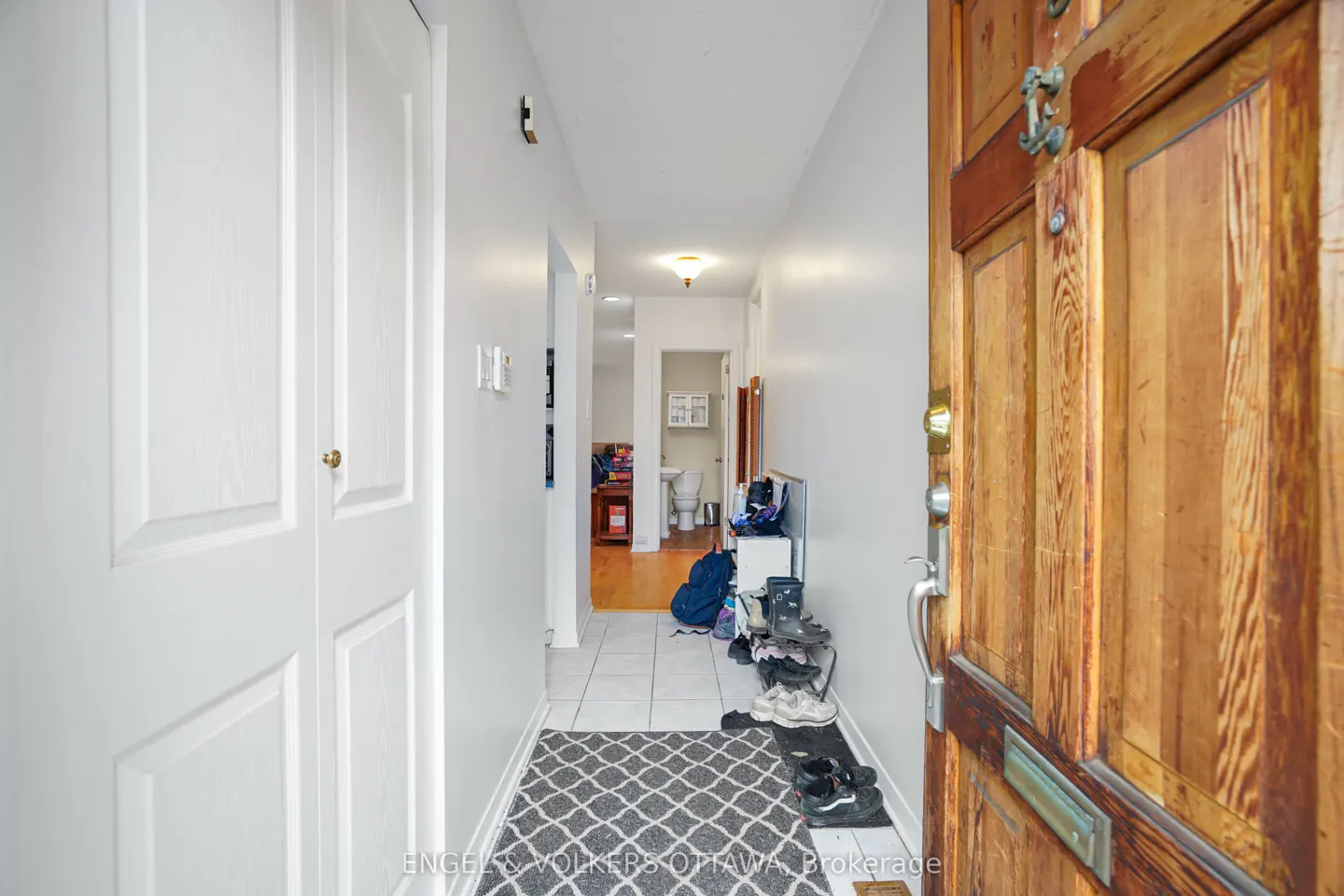 Entryway with a wood door, white walls, and a gray patterned rug. Shoes and a backpack are near a doorway leading to a bathroom.