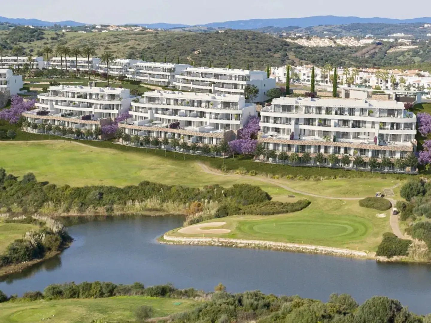 Aerial view of white modern apartment buildings overlooking a green golf course with a lake and rolling hills in the distance.