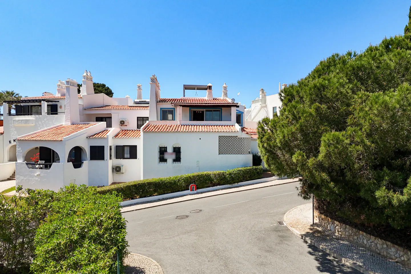 Exterior view of white villas with red tile roofs under a clear blue sky, beside a road with green trees.
