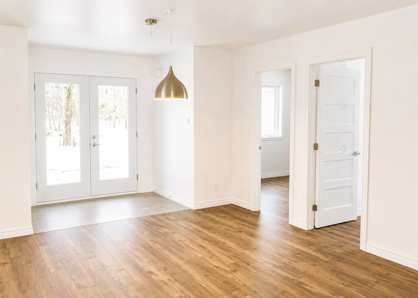 Bright, empty room with wood floors, white walls, and white trim. French doors lead to a snowy yard. A gold pendant light hangs from the ceiling.