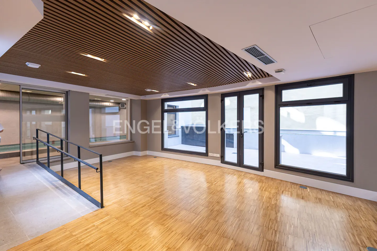 An empty room with wood floors, gray walls, and a slatted wood ceiling. Black framed windows and doors look out onto a patio.