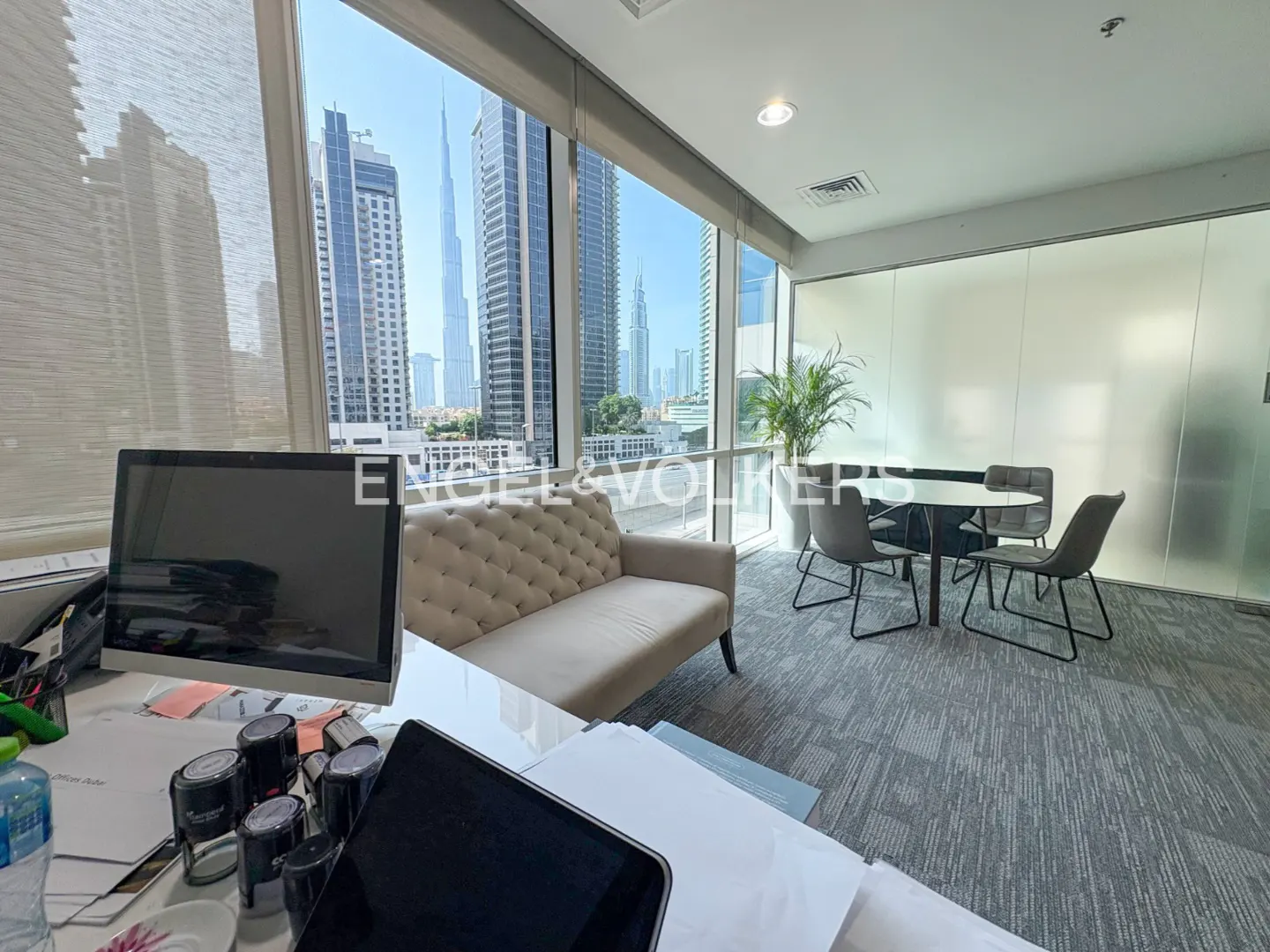 Office space with a beige sofa, round table with chairs, and a view of Dubai skyscrapers through large windows.