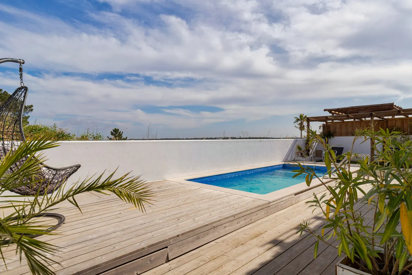 Outdoor patio with a blue pool, wooden deck, white wall, and hanging chair under a cloudy sky.
