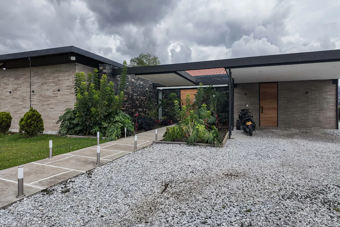 Modern home exterior with stone driveway, walkway, and lush landscaping. A motorcycle is parked near the wooden front door. Overcast sky.