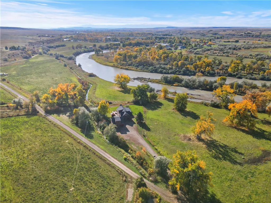 Aerial view of a dark house on a green lawn with fall foliage, near a winding river under a blue sky.