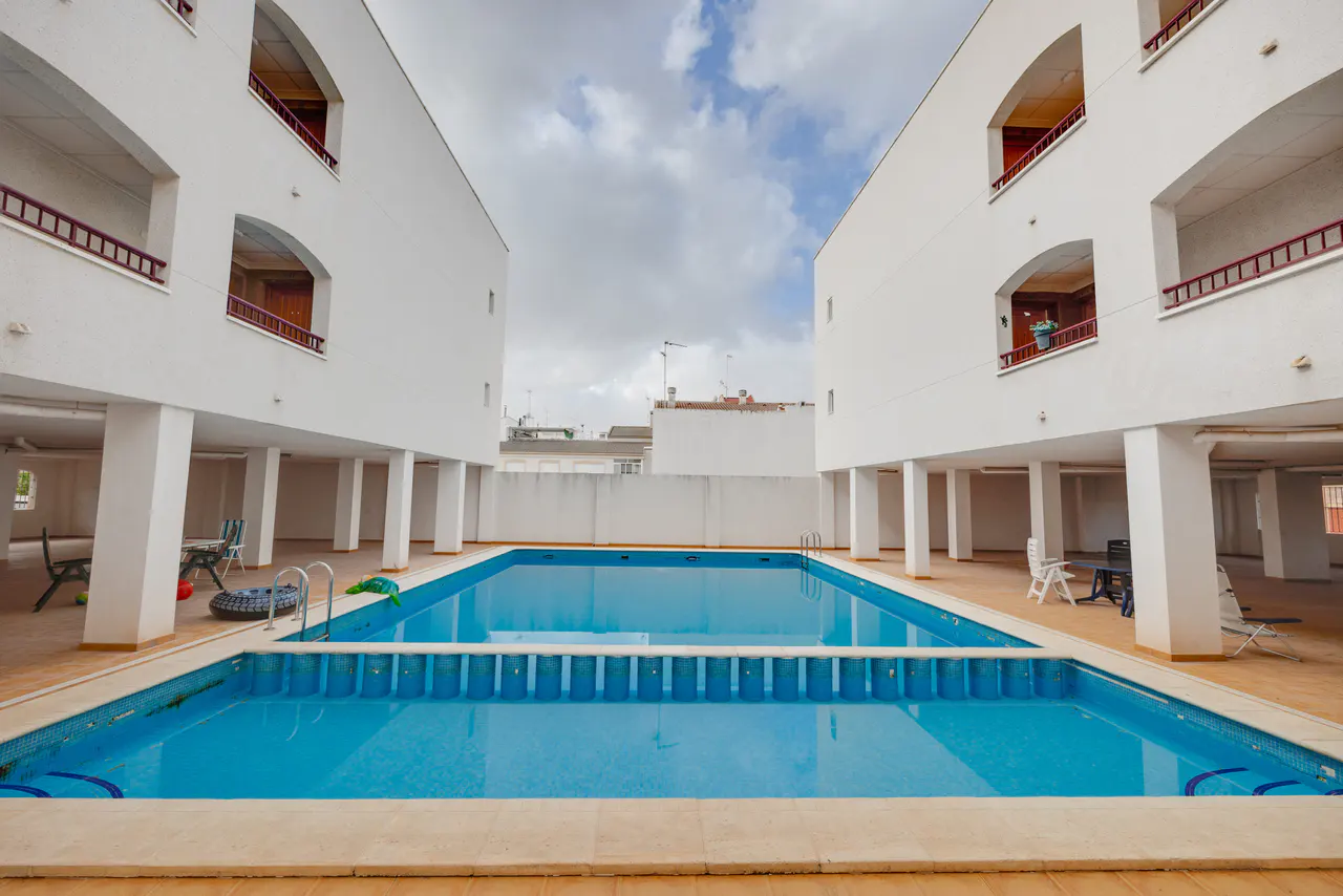Rectangular blue swimming pool between two white apartment buildings with balconies and support columns.