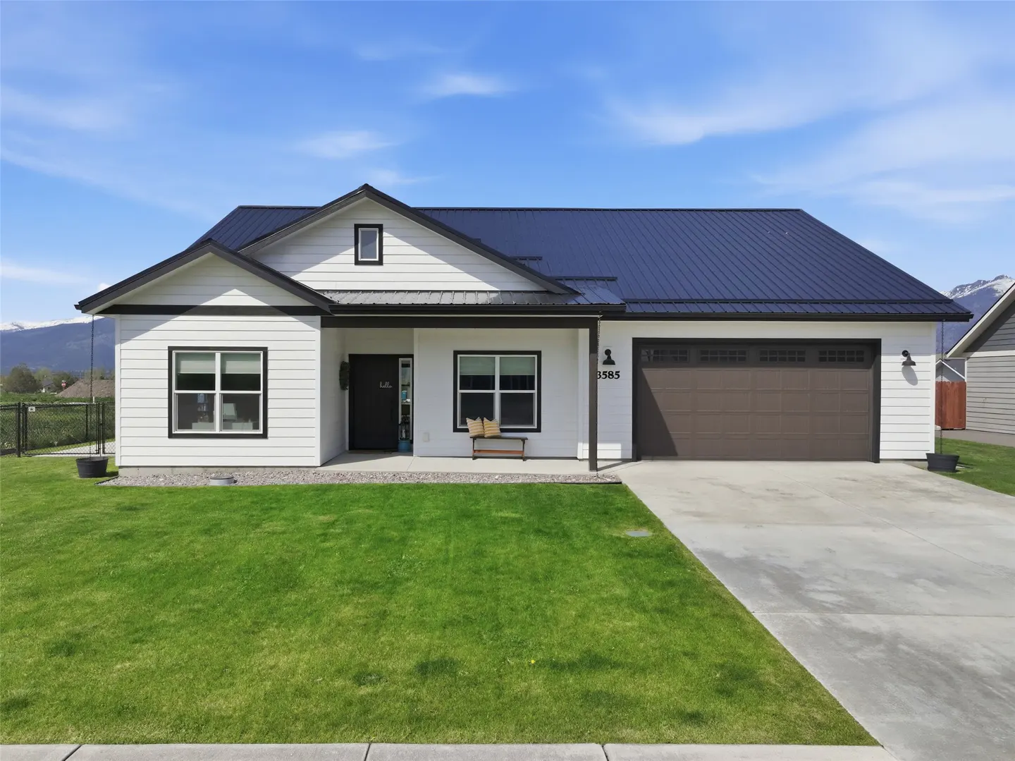 A white, one-story house with a dark metal roof and a brown garage door sits on a green lawn under a blue sky.