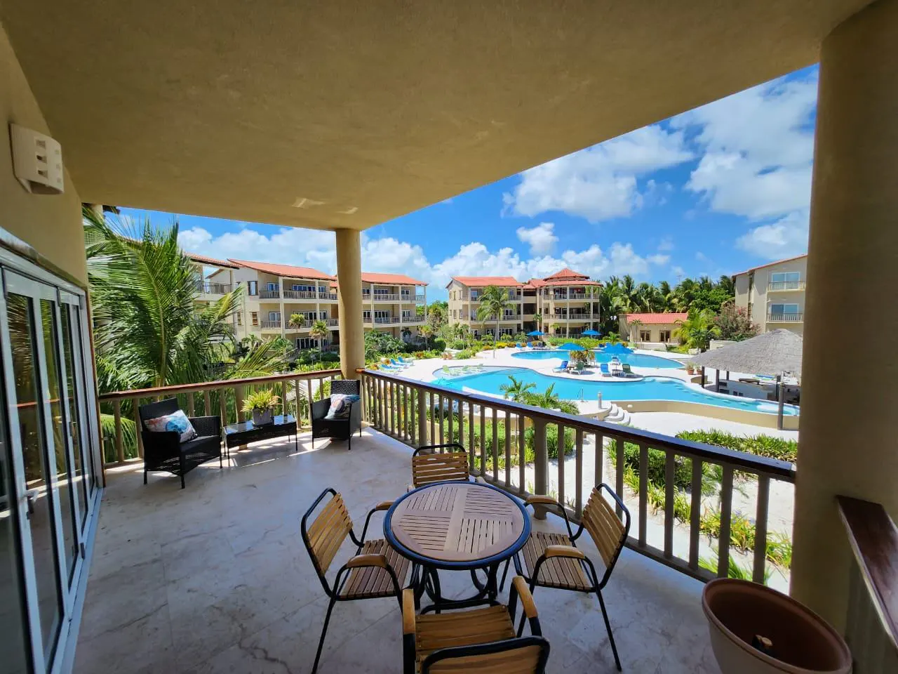 Balcony view of a resort with a pool. Table and chairs on the balcony, with lounge seating to the left. Buildings and palm trees in the background.