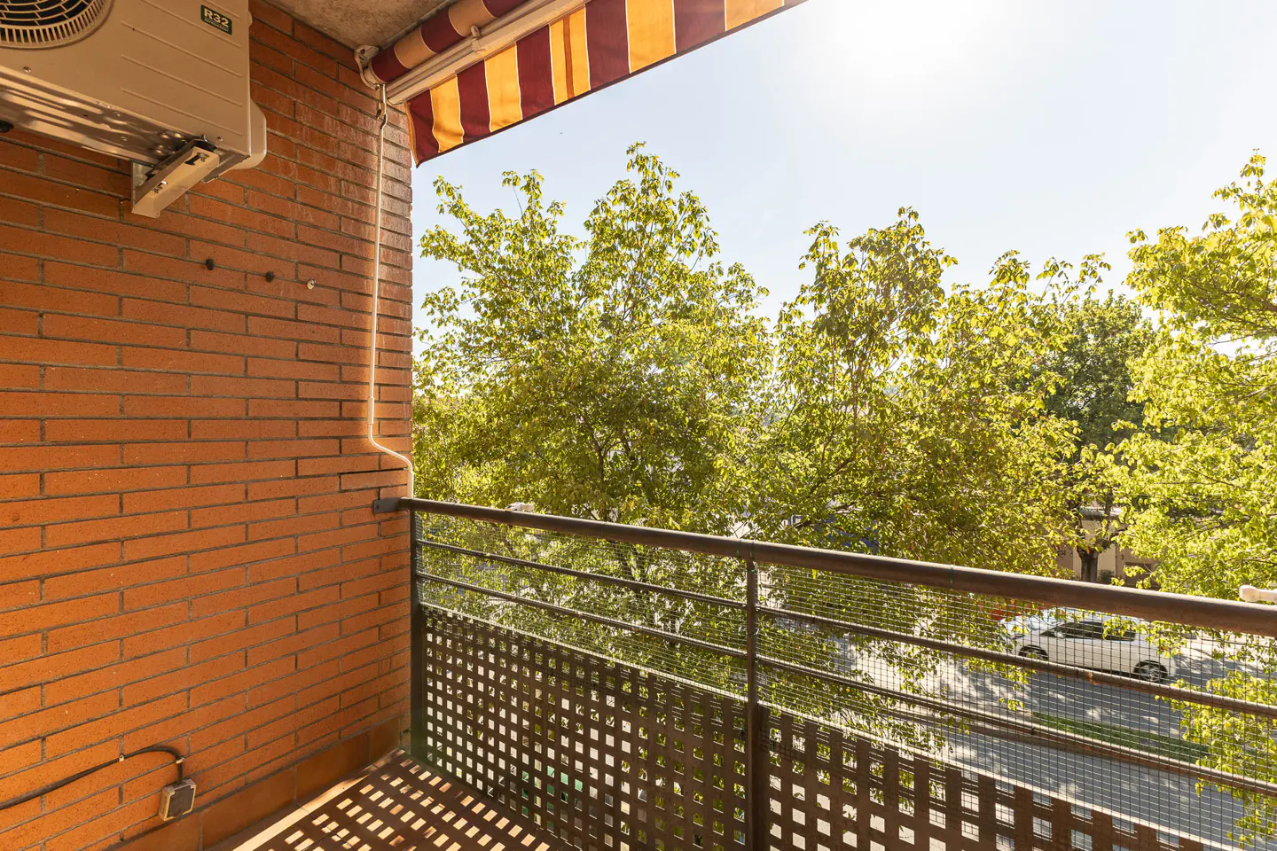 Balcony view with brick wall, metal railing, and striped awning. Green trees and a street with a car are visible in the background.