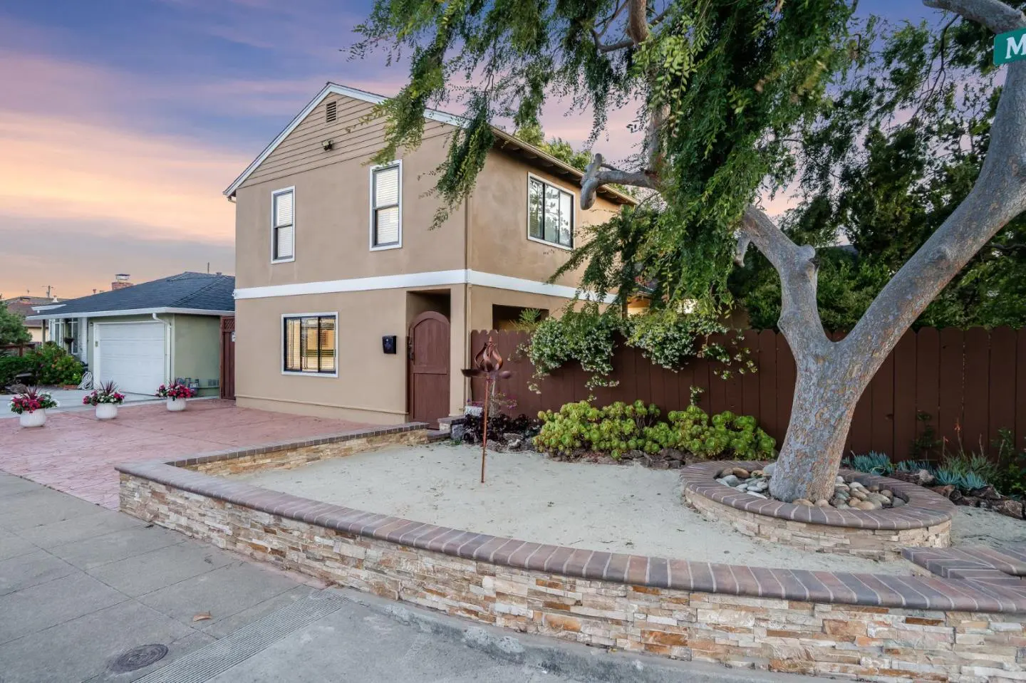 Two-story tan house with a brown door and black-framed windows. A tree is in the front yard, surrounded by rocks and a stone wall.