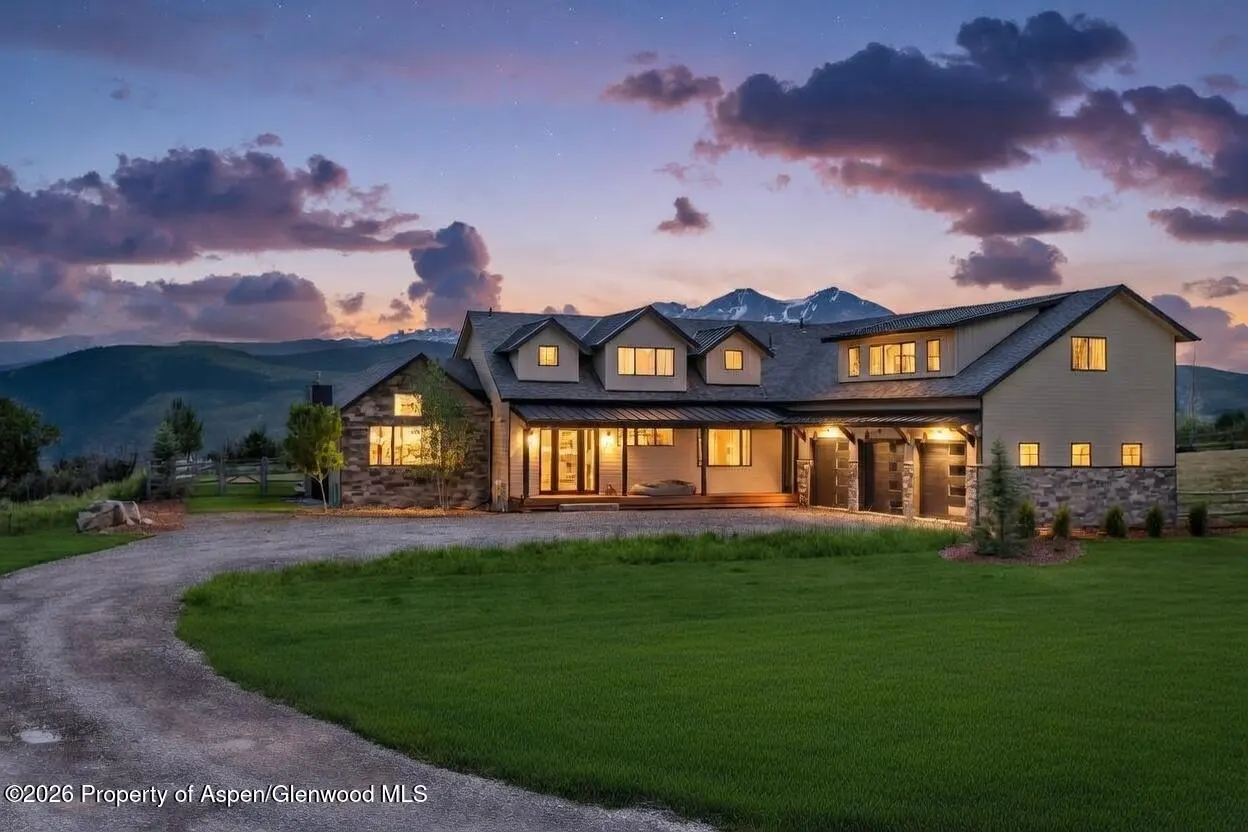 A large, two-story house with stone and siding exterior, set against a mountain backdrop at dusk. The sky is a mix of purple and orange hues.