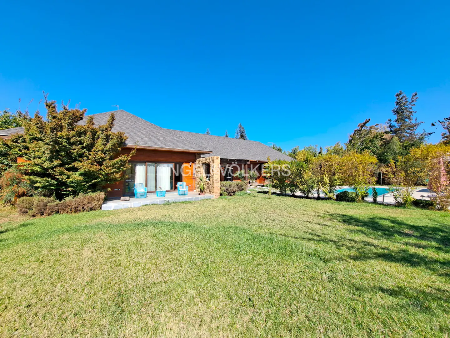 Exterior view of a single-story home with a green lawn, trees, and a blue sky. A pool is visible in the background.