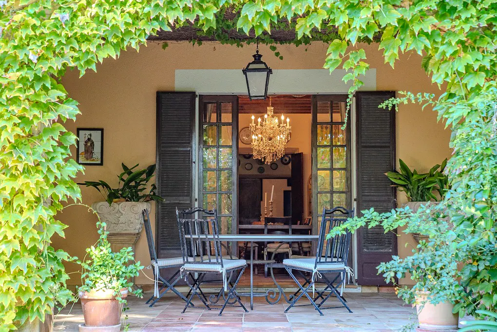 Outdoor patio with a table and chairs, framed by green vines. Open doors reveal a chandelier inside.