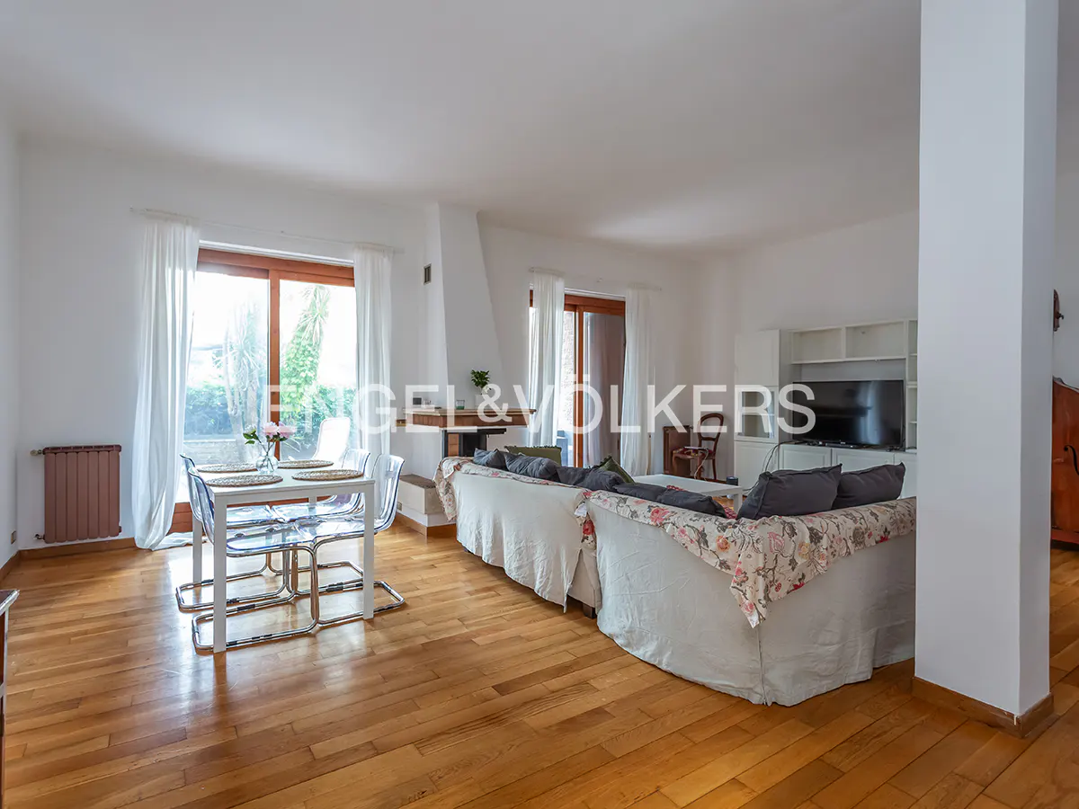 Bright, open-plan living room with wooden floors, white walls, dining table with clear chairs, and a large sofa with floral accents.