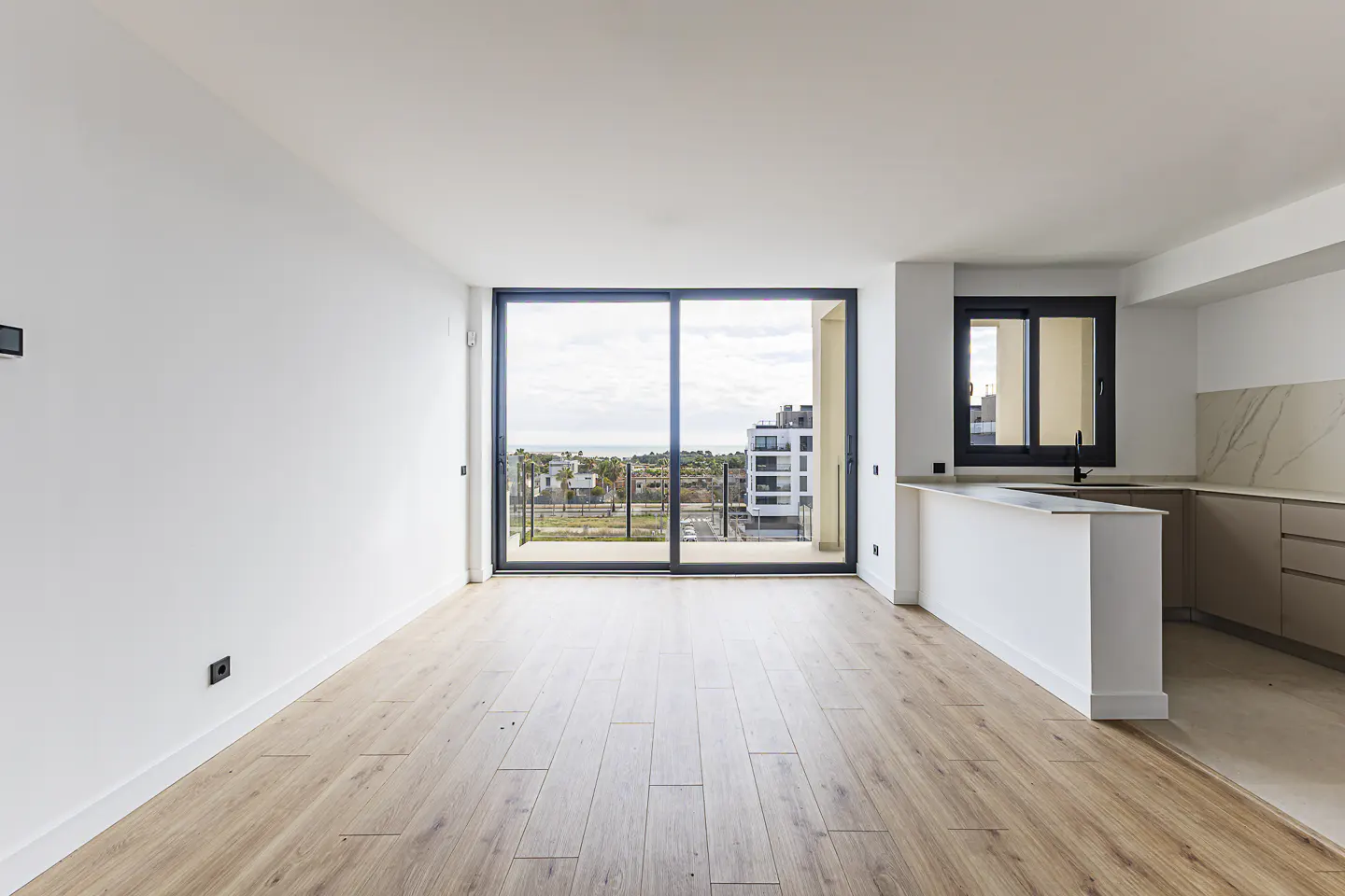 Bright, modern apartment interior with wood floors, white walls, and a sliding glass door to a balcony with a city view. Kitchen area visible.