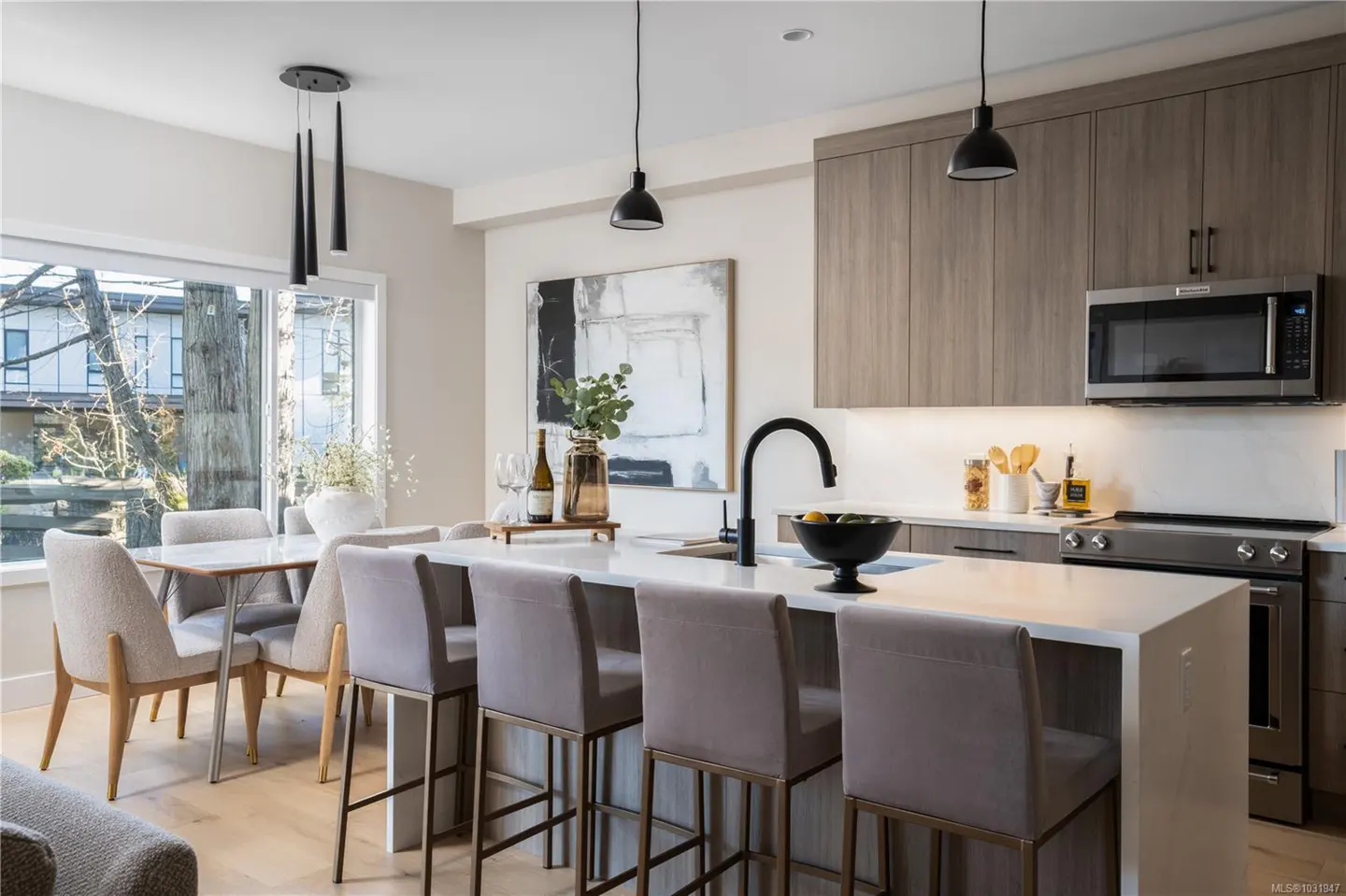 A modern kitchen with a white island, gray cabinets, and black pendant lights. A dining table sits near a window with a view of trees.
