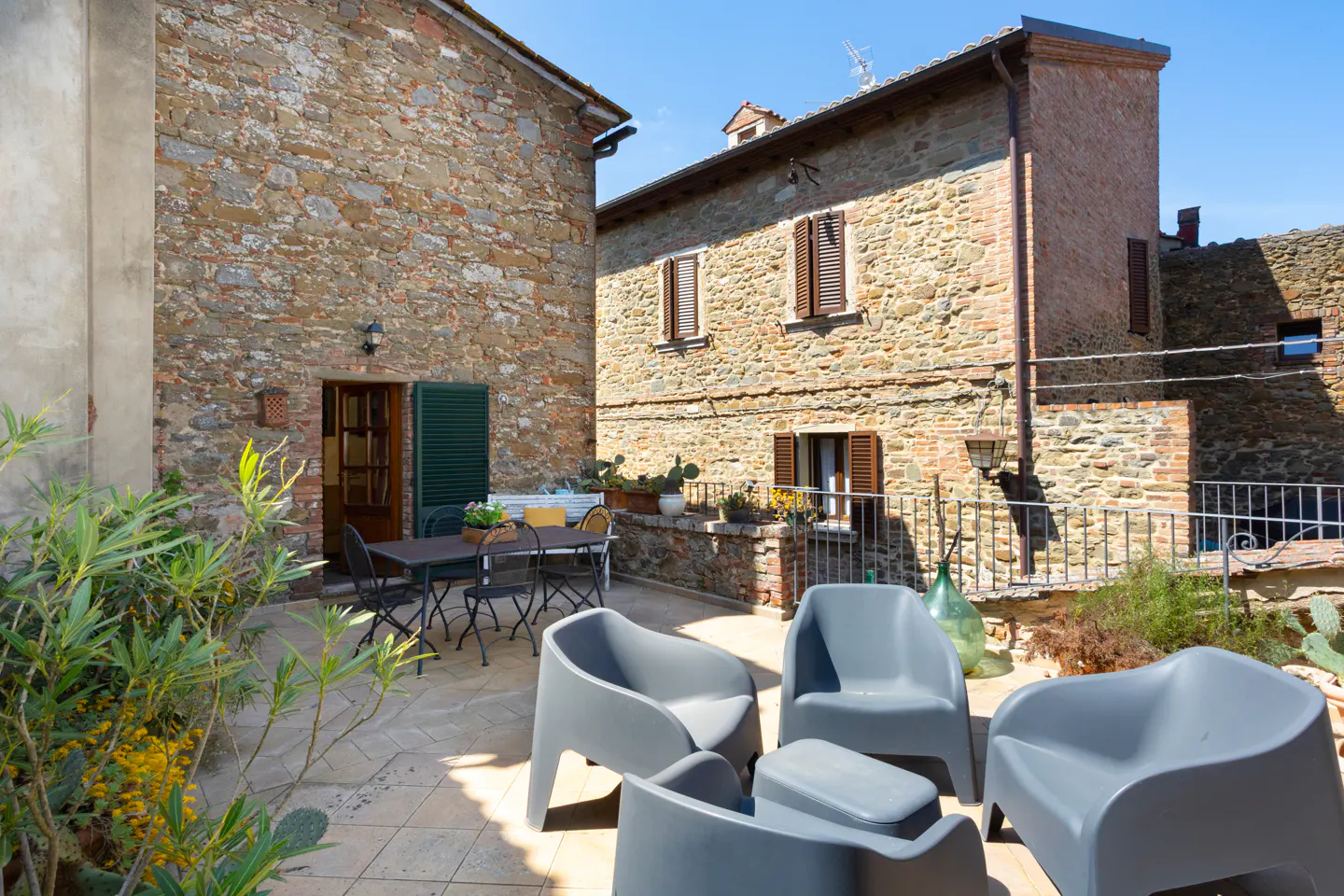 Outdoor patio with gray chairs and table, stone buildings in background, green plants, blue sky.