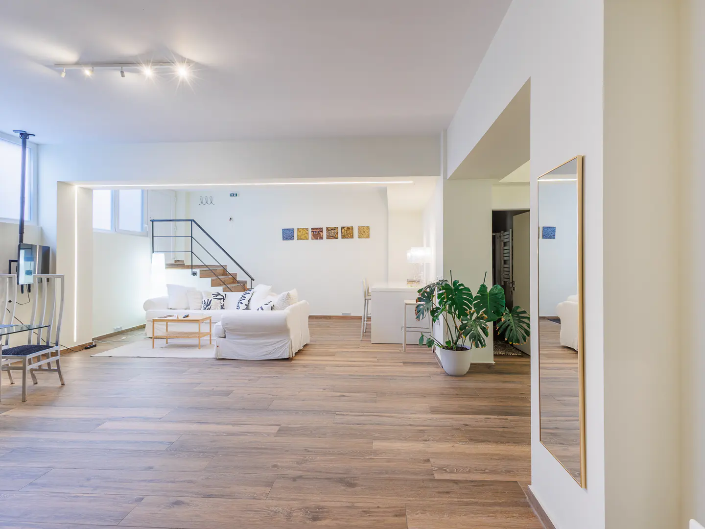 Bright, open-concept living space with hardwood floors, white walls, and a staircase. A white sofa and potted plant add warmth.