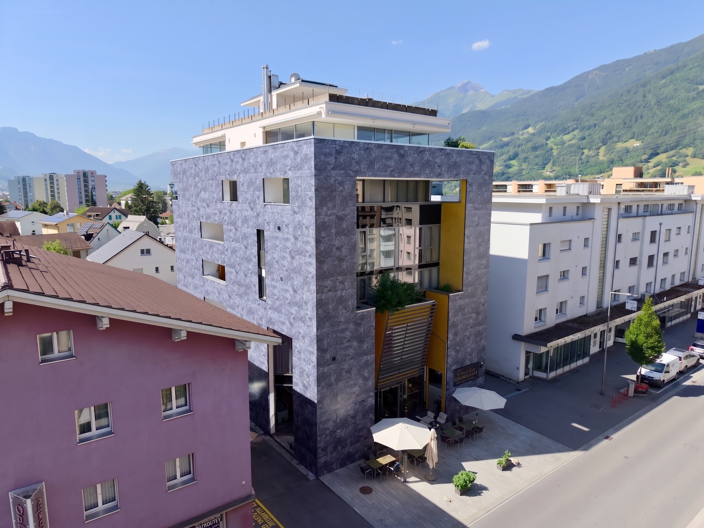 Modern grey stone building with a yellow accent wall and outdoor patio seating, set against a backdrop of mountain peaks.