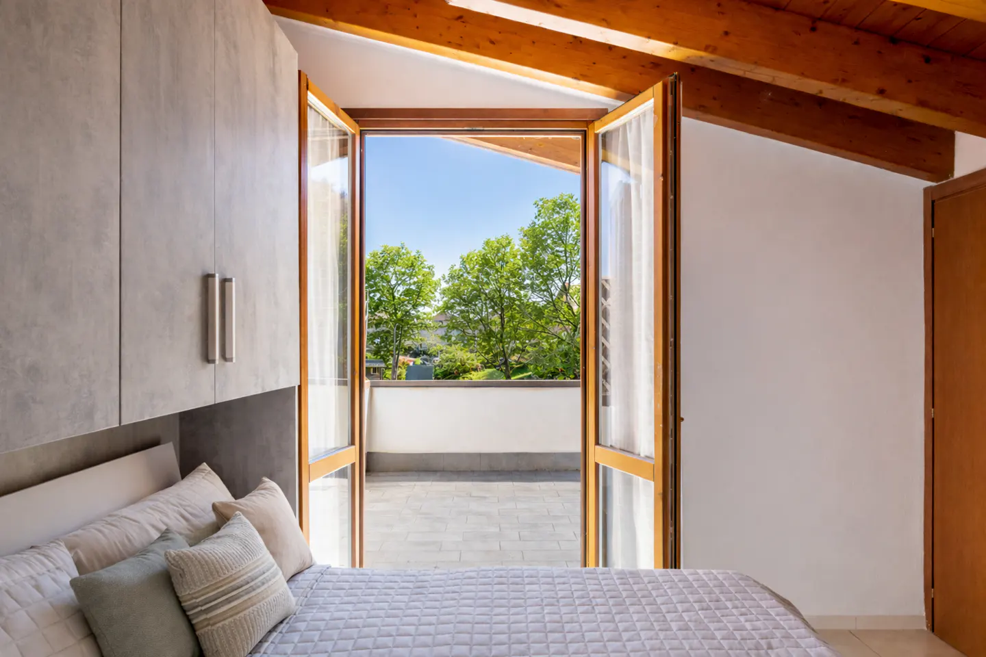 Bedroom with a gray quilted bed, pillows, and open doors to a balcony with trees and blue sky.