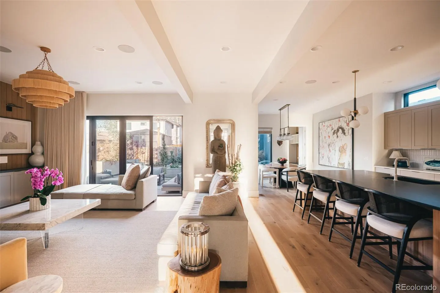 Bright, open-concept living space with sofas, a kitchen island with stools, and a dining area. A Buddha statue is centered on the wall.