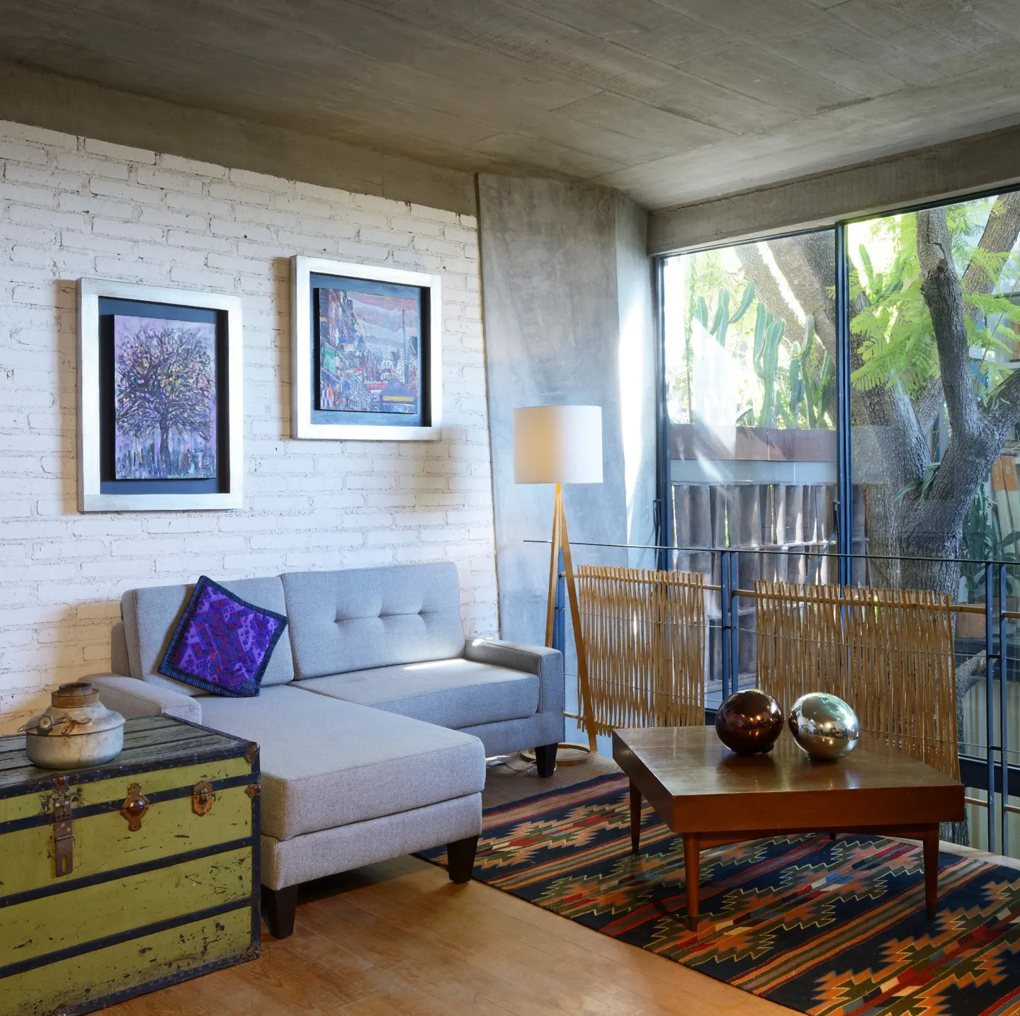 Living room with gray sofa, purple pillow, and green trunk. Art hangs on the white brick wall. A large window shows trees outside.