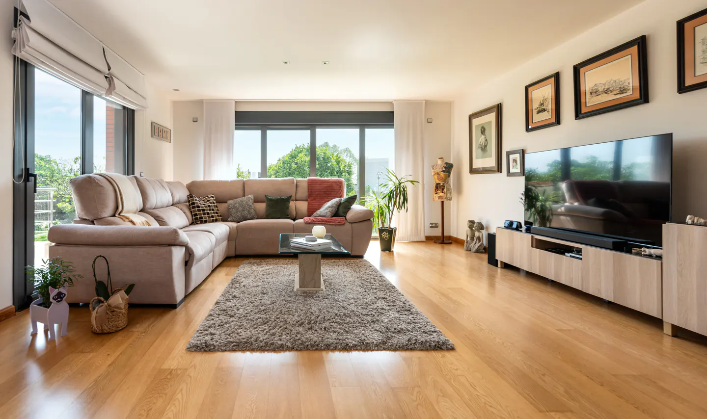Living room with a beige sectional sofa, a gray rug, and a TV on a light wood stand. Large windows offer a view of greenery.