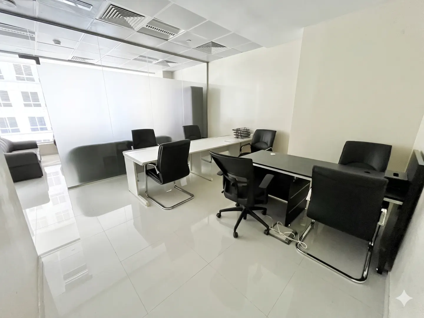 Bright office space with white walls and floors, featuring black leather chairs and desks. A gray sofa sits behind a glass partition.