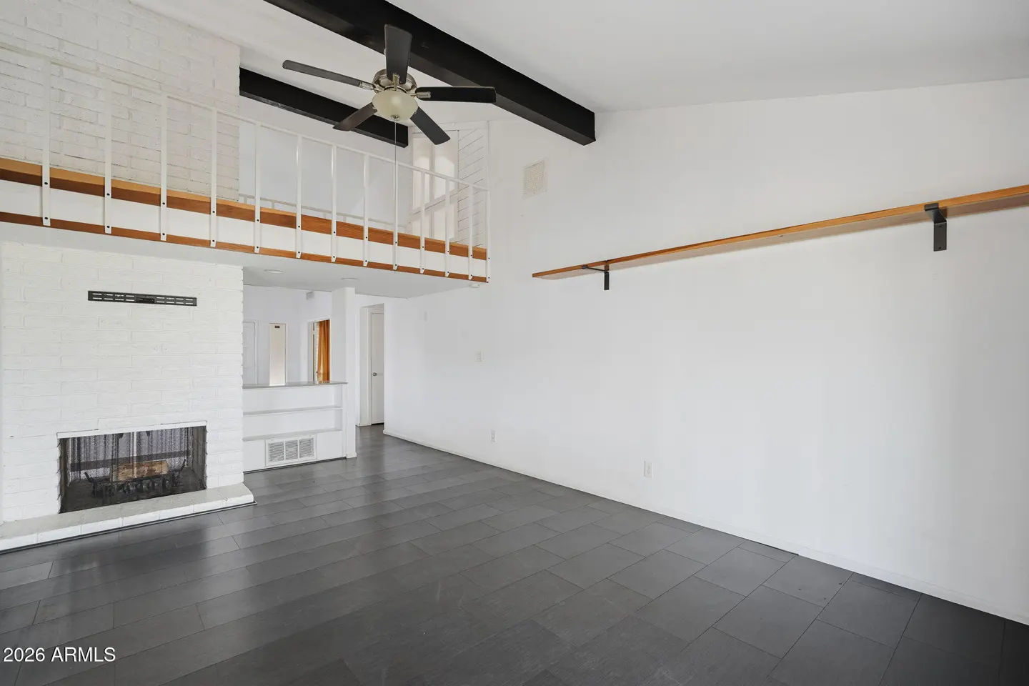 A bright living room with a white brick fireplace, black ceiling beams, and dark gray tile floors. A white railing overlooks the room.