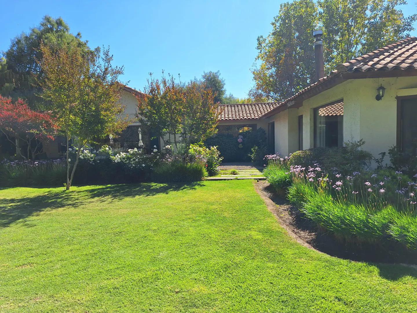 Lush green lawn leads to a white house with a red tile roof. Trees and flowers surround the house. Blue sky.