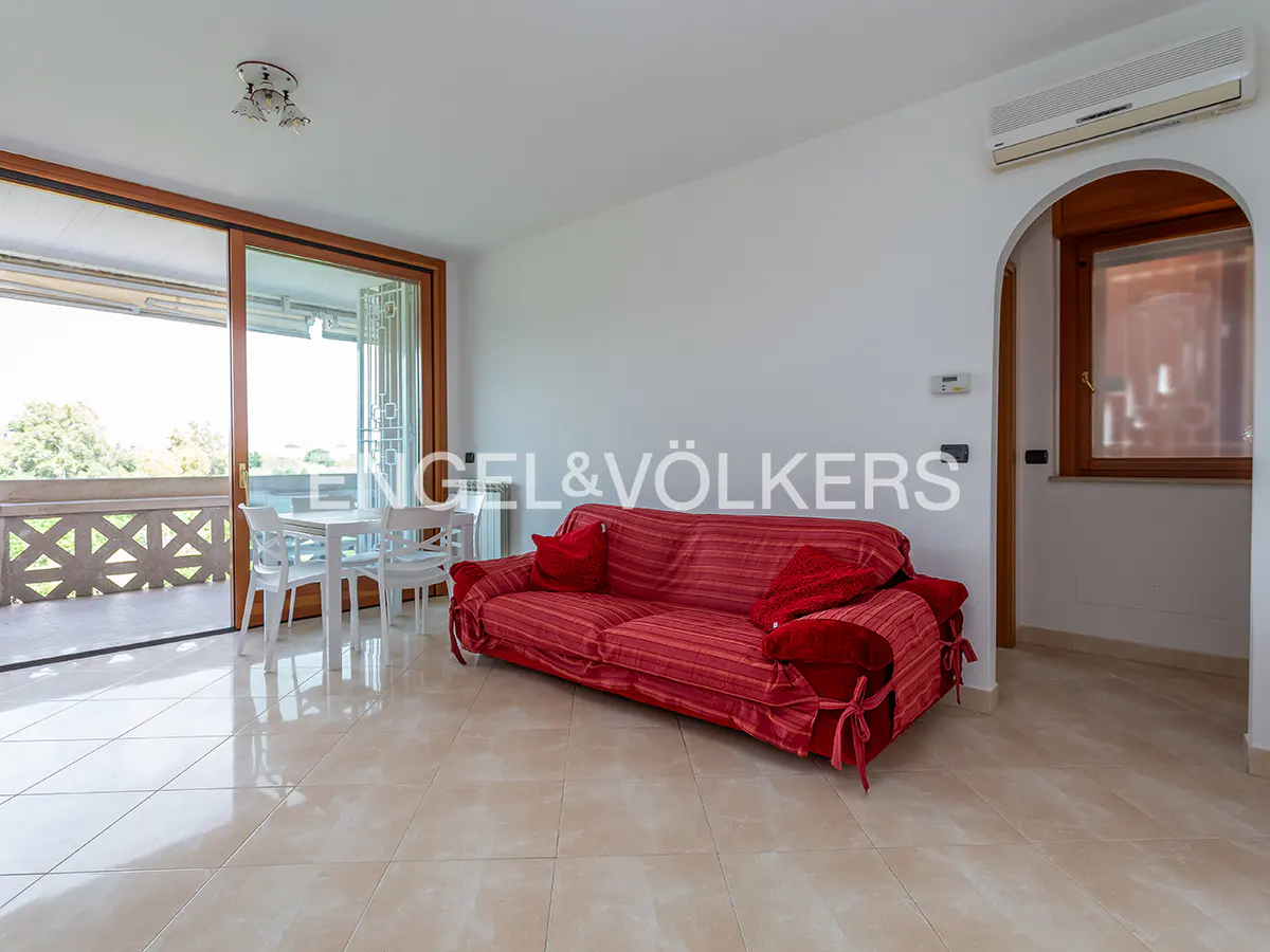 Bright living room with a red sofa, white table and chairs, and a balcony with a view. Tiled floor and white walls.
