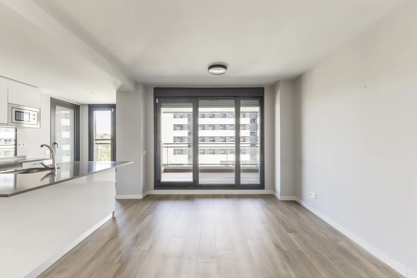 Bright, modern apartment interior with wood floors, white walls, and a kitchen island. Sliding glass doors lead to a balcony with a city view.