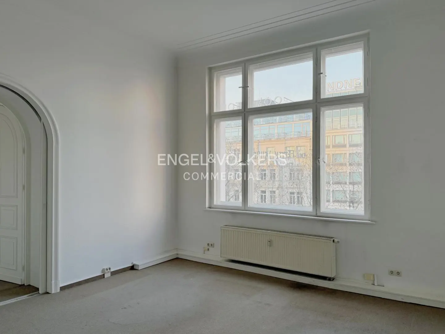 Bright, empty room with white walls, a large window showing a city view, and a radiator. An arched doorway is visible on the left.