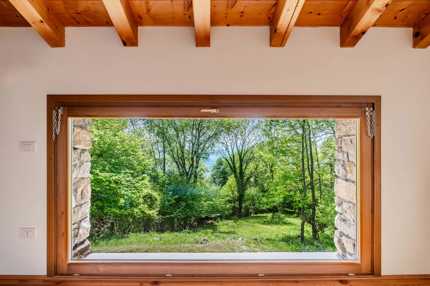 A window with a wood frame and stone sides looks out onto a green forest. The ceiling has exposed wood beams.